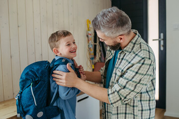 Father helping son get ready for kindergarten, preschool. Putting backpack on back.