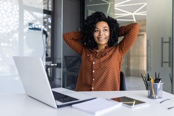 Obraz premium A young African American businesswoman with natural curly hair, dressed in a stylish polka dot blouse, relaxes at her workspace filled with light, exuding confidence and comfort.