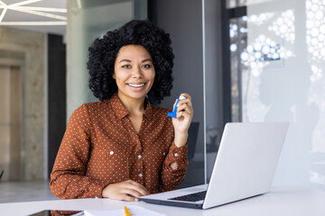 African American businesswoman using a blue asthma inhaler while working on her laptop at a stylish office setting. She seems relieved and focused.