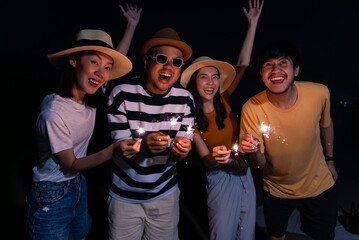 Cheerful asian diverse group friends are dancing and waving sparklers enjoying warm summer nights outdoors on beach. Youth and leisure activities concept.