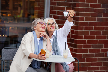Sitting Outdoor Cafe stylish mature women with smiles capturing joyful selfie moment together, wearing glasses and smart casual attire, with drinks on table, relaxed social outing of elderly people