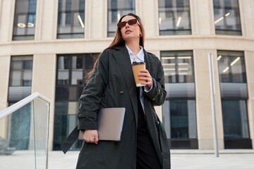 Confident young Caucasian woman with black hair walks through the city with a laptop in her hand