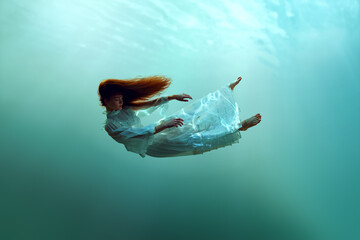 Creative image depicting redhead young calm girl levitating underwater showcasing the harmony of humans and nature. Concept of surrealism, beauty, mystery and fantasy, freedom