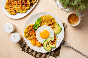 Homemade breakfast or brunch. Fried egg with belgian waffles made with cheese and vegetables, zucchini, onion, carrot, served with avocado and yellow tomato. Cup of coffee drink.