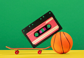 Creative still life with audio tape, skateboard and basketball ball on green and yellow background.