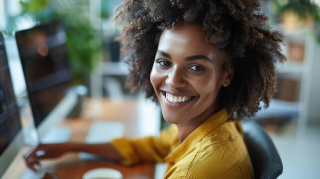 Smiling woman with curly hair wearing yellow top sitting at desk with com puter monitor and keyboard in modern office environment with blurred greenery in background.