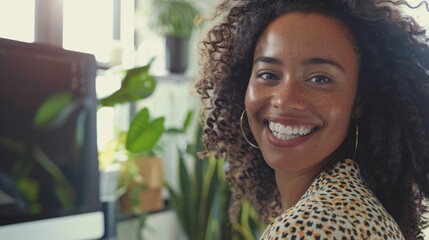 Smiling woman with curly hair and freckles wearing a leopard print top standing in a brightly lit room with plants and a monitor in the background.