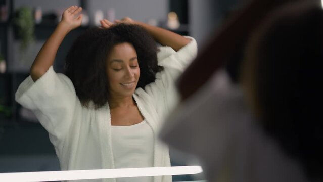 African American happy pretty woman looking mirror reflection morning biracial ethnic girl lady female at bathroom home bath stretch yawn pampering check hairdo touch face hair beauty routine wake up
