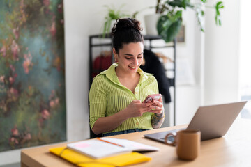 A content Hispanic woman in a green-striped shirt enjoys her smartphone at a modern workspace, reflecting a seamless blend of technology and lifestyle