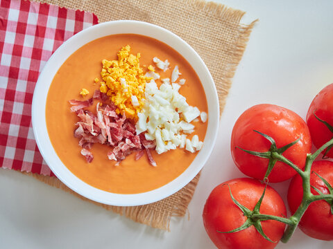 Spanish cold tomato soup called salmorejo. Served in a bowl, with chopped hard-boiled egg and diced Serrano ham. Fresh red tomatoes on the table. Close-up view from above.