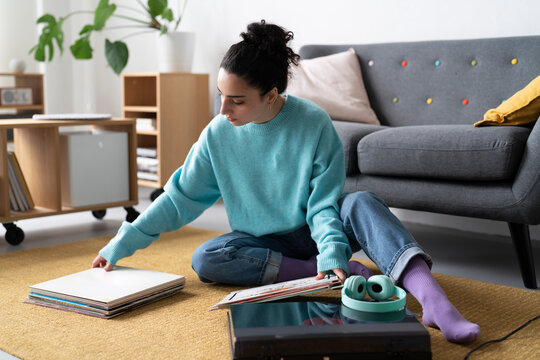 A young woman in a blue sweater examines a vinyl record, engaged in her passion for music, with a turntable and headphones nearby