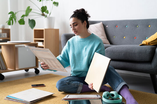 A young woman in a blue sweater examines a vinyl record, engaged in her passion for music, with a turntable and headphones nearby