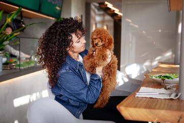 Portrait of a beautiful curly brunette woman cuddling her poodle pet in a restaurant. Elegant female with her small dog enjoying indoor dining