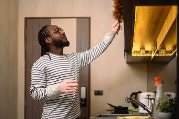 Happy bearded African man dusting shelf with a feather duster at modern kitchen