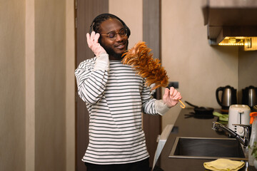 Young African American man wearing headphone doing chores and singing with a feather duster