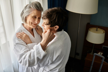 A senior lesbian couple in bathrobes sharing a tender embrace in a hotel.