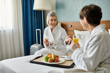 Senior lesbian couple in bathrobes enjoys a tray of fruit on the bed.