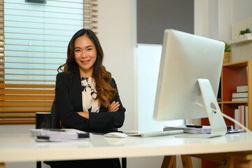 Portrait of confident female manager in elegant suit sitting at her workplace and smiling to camera