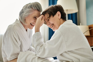 Two senior women share a moment of laughter on a cozy bed.