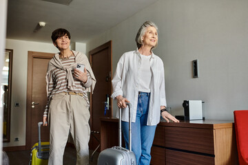Senior lesbian couple stands with luggage in hotel room.