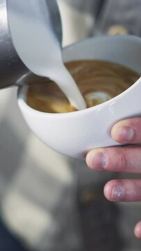 Professional barista pouring milk foam over coffee into white cup creating perfect cappuccino and making a nice pattern. Vertical video.