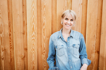 Smiling Blonde Woman in Denim Shirt Standing Outdoors Against Wooden Background