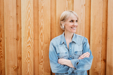 Laughing Blonde Woman in Denim Shirt with Folded Arms Outdoors against Wooden Wall
