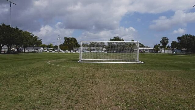 A drone video shows an aerial view of a green soccer field in a park. People walk and exercise while the houses of a peaceful neighborhood as a backdrop. Open space for outdoor activities and sports.
