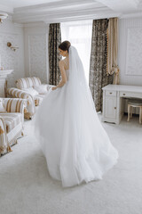 Fototapeta na wymiar A woman in a white wedding dress is standing in front of a desk. The room is decorated with a lot of furniture, including two chairs and a desk. The woman is wearing a veil