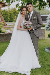 A bride and groom are posing for a picture in a garden. The bride is wearing a white dress and the groom is wearing a suit. They are holding a bouquet and a bouquet of flowers