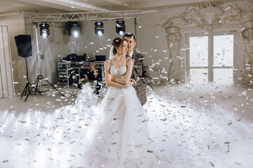 A bride and groom are dancing in a room with confetti falling around them. Scene is joyful and...