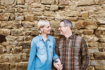 A happy couple, a 50-year-old man and a 40-year-old woman, stand outside in front of a stone wall, smiling and gazing into each other's eyes