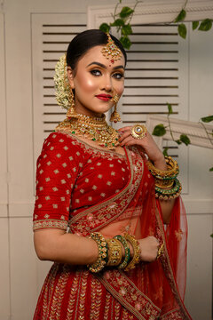Stunning Indian bride adorned in a traditional red bridal lehenga gracefully wears heavy gold jewellery and a veil and smiles tenderly in studio lighting. Wedding fashion and lifestyle.