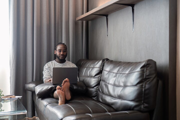 Young African American man holding laptop relaxing sitting on sofa working remote at home