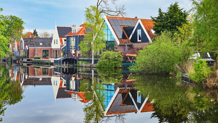 A picturesque scene showing a row of charming houses nestled next to a tranquil body of water, reflecting the clear blue sky and lush greenery