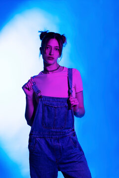 Beautiful young woman in bib overalls standing against illuminated background