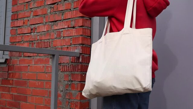 Woman in red hoodie holding tote canvas blank eco bag on street brick wall background. Female consumer hold white textile shopper. template or place for your design, logo, text