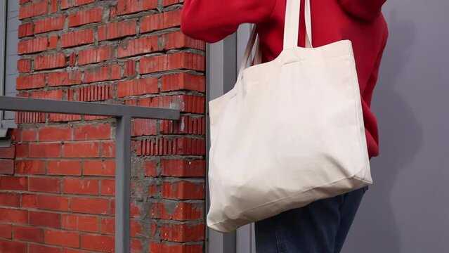 Woman in red hoodie holding tote canvas blank eco bag on street brick wall background. Female consumer hold white textile shopper. template or place for your design, logo, text
