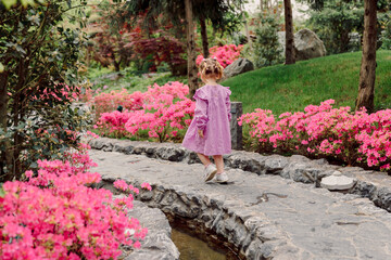 Child girl in stylish dress walking in summer garden with pink flowers.