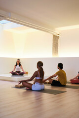 A group of people are meditating, sitting on yoga mats in a room. Scene is peaceful and serene, as the people are practicing yoga.