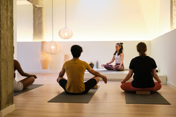 A group of people are sitting on yoga mats meditating in a room. One woman is teaching the others how to meditate.