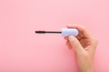 Young adult woman hand holding and showing purple tube of black eyes mascara on light pink table background. Pastel color. Female beauty product. Closeup. Top down view. - 800102364