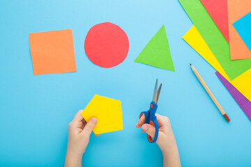 Little child hands holding scissors and cutting colorful geometric shapes from application paper on pastel blue table background. Making different forms. Point of view shot. Closeup. Top down view. - 800102385