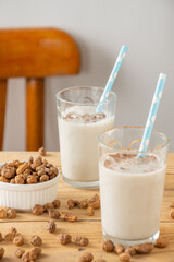 Close-up of two glasses with horchata and straws on wooden table with tiger nuts, white background with chair, vertical, with copy space