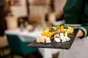 Person in green attire holding a black slate tray with assorted cheeses, fruits, and honey, set against an elegant dining backdrop