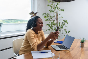 Happy woman wearing headphones sitting with laptop at table in home office. Successful African...