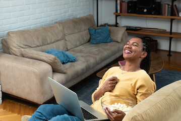 Cheerful young black woman with dreadlocks watching comedy movie on laptop at home, sitting on...
