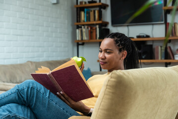 Simple living. African American woman reading book and eating apple in living room. Healthy life...