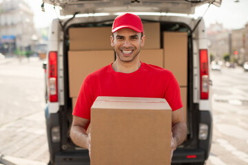 Happy young courier man in red uniform standing with parcel box near car outdoors and smiling at...