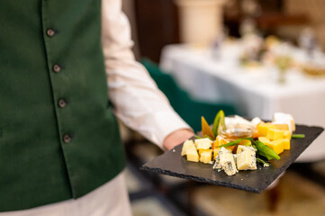 Waiter in green vest and white shirt, presenting a slate tray with assorted gourmet cheeses, embodying fine dining elegance